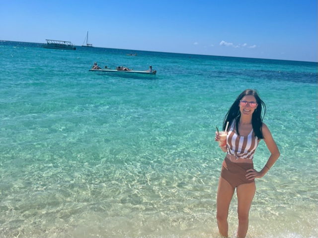 Girl in brown swimming suit standing on beach near turquoise water.