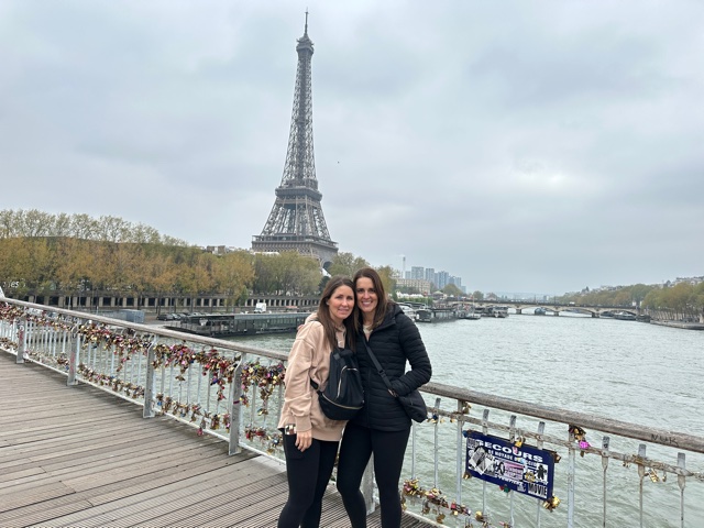 Two woman standing on a bridge with tower behind them.